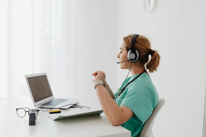 A nurse wearing headphones is sitting at a desk in front of a laptop computer taking enquiries from customers.
