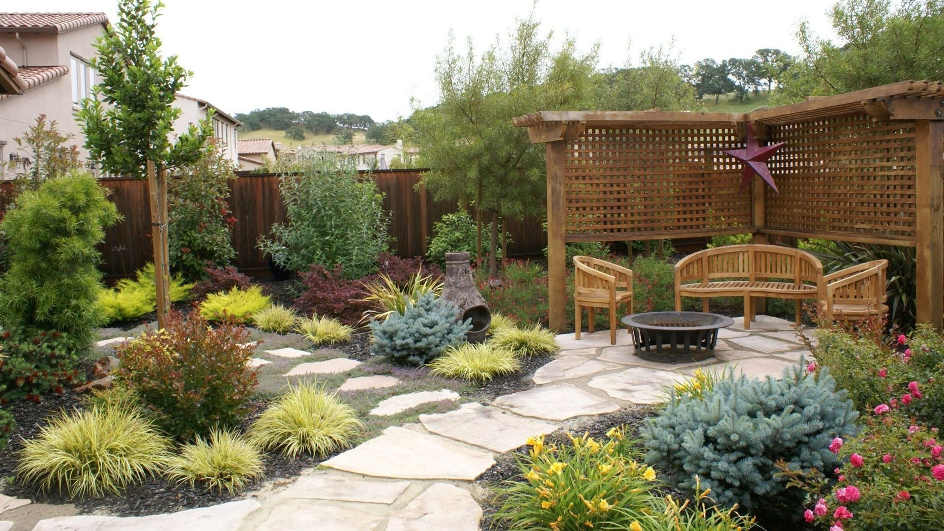 Cozy backyard landscape design with lush plants, a stone pathway, and a wooden pergola with seating and a fire pit.
