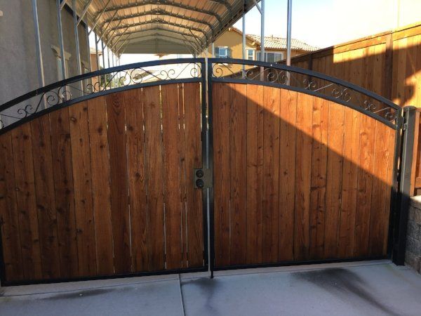 A wooden driveway gate with decorative wrought iron accents, set beneath a covered carport and surrounded by a wooden fence.