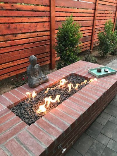 A rectangular brick fire pit with flames over lava rocks, with a Buddha statue and a tray with cups.