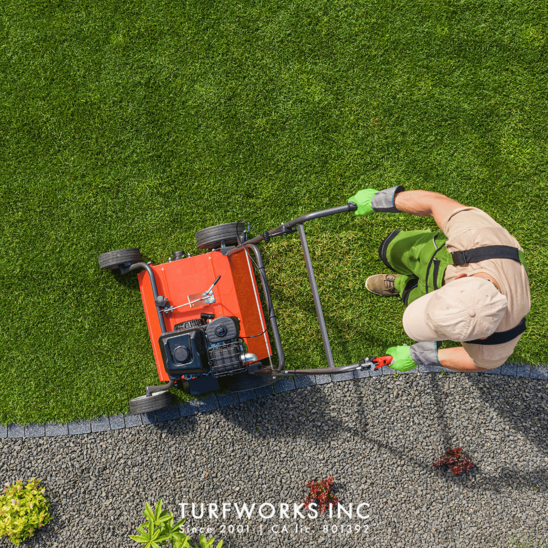 Overhead view of a landscaper in green gloves and a tan cap running a lawn dethatcher along a gravel-bordered lawn.