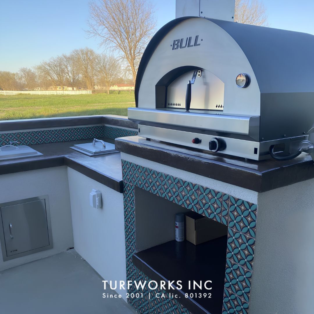 Bull pizza oven on decorative tiled outdoor kitchen base, overlaid with Turfworks Inc logo.