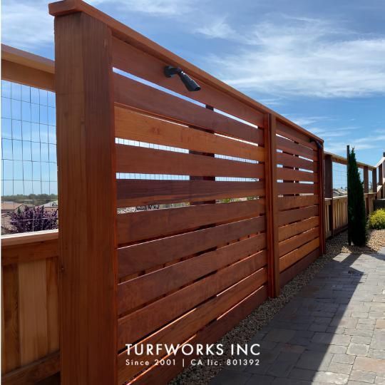 Horizontal wood slat privacy fence with mounted spotlight and paver pathway alongside, with Turfworks Inc logo.