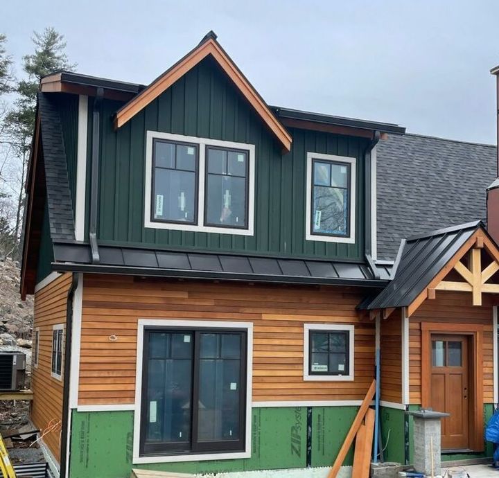 Two-story house under construction with green and brown siding, dark roof, and windows.