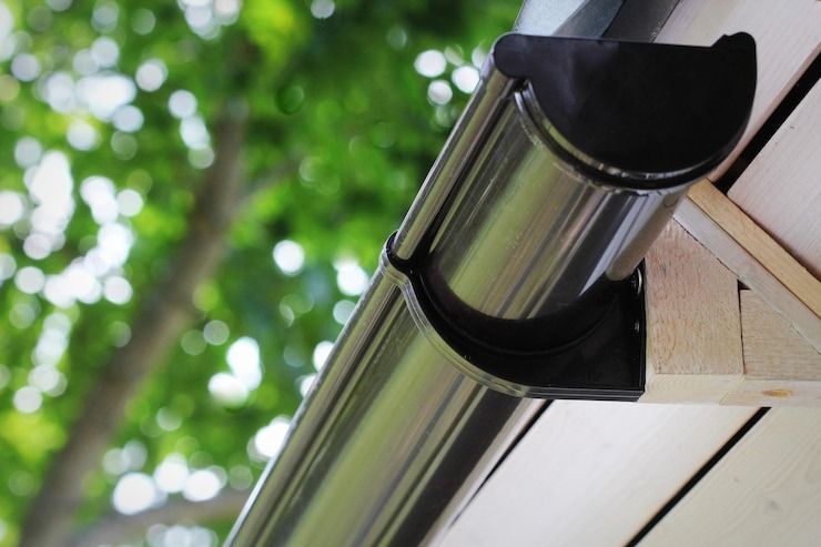 Dark brown rain gutter attached to a white wooden house trim, with a blurred green tree background.