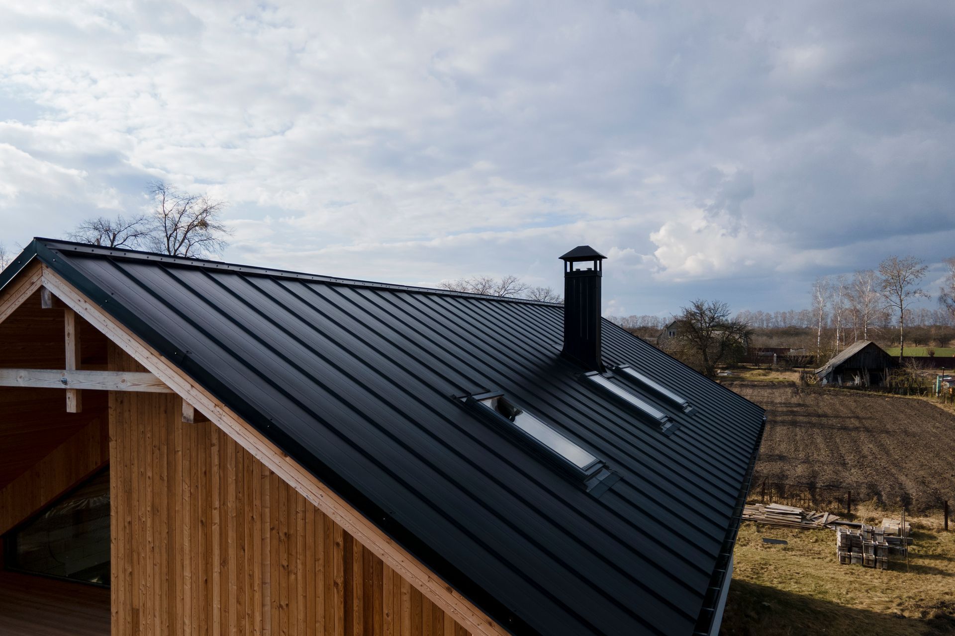 Black metal roof on a wooden structure with a chimney and skylights, set in a rural area.
