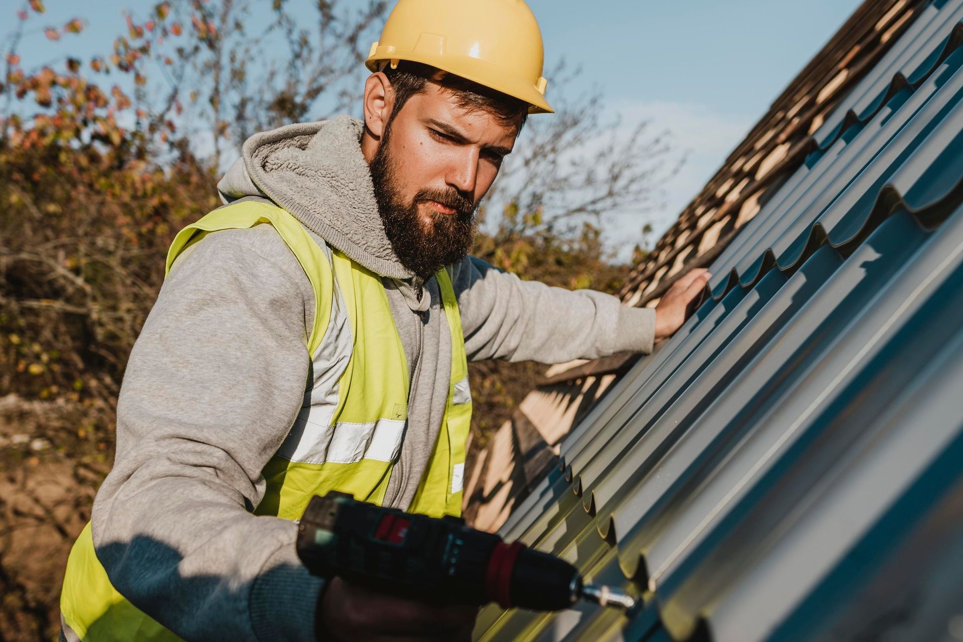 Construction worker in hard hat and safety vest installing metal roofing with a power drill.