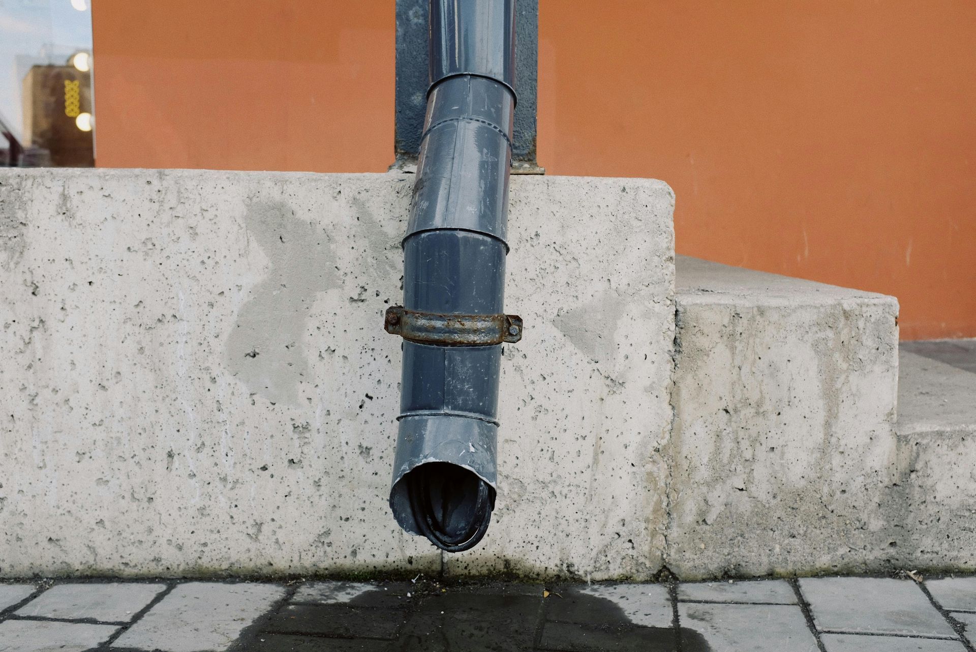 Gray rain gutter against concrete steps and orange wall, water dripping from the end.