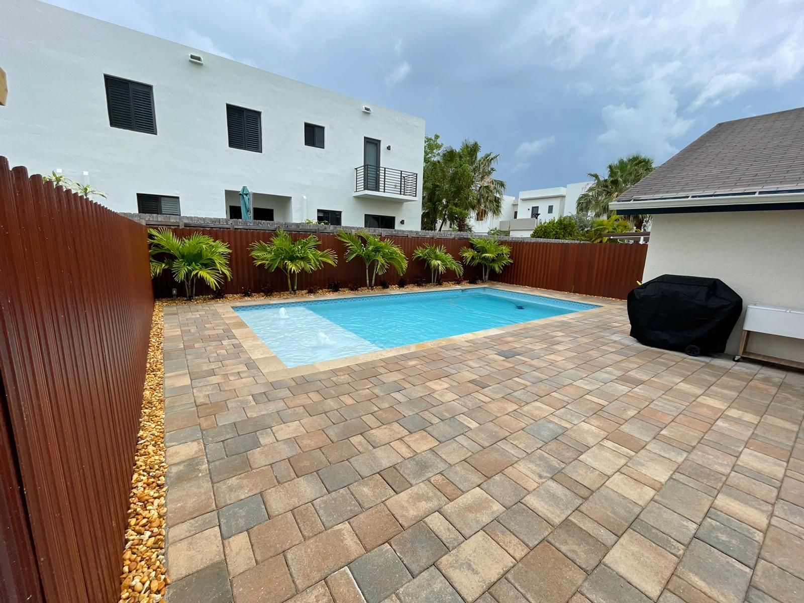 A large swimming pool is surrounded by a brick patio and a wooden fence.