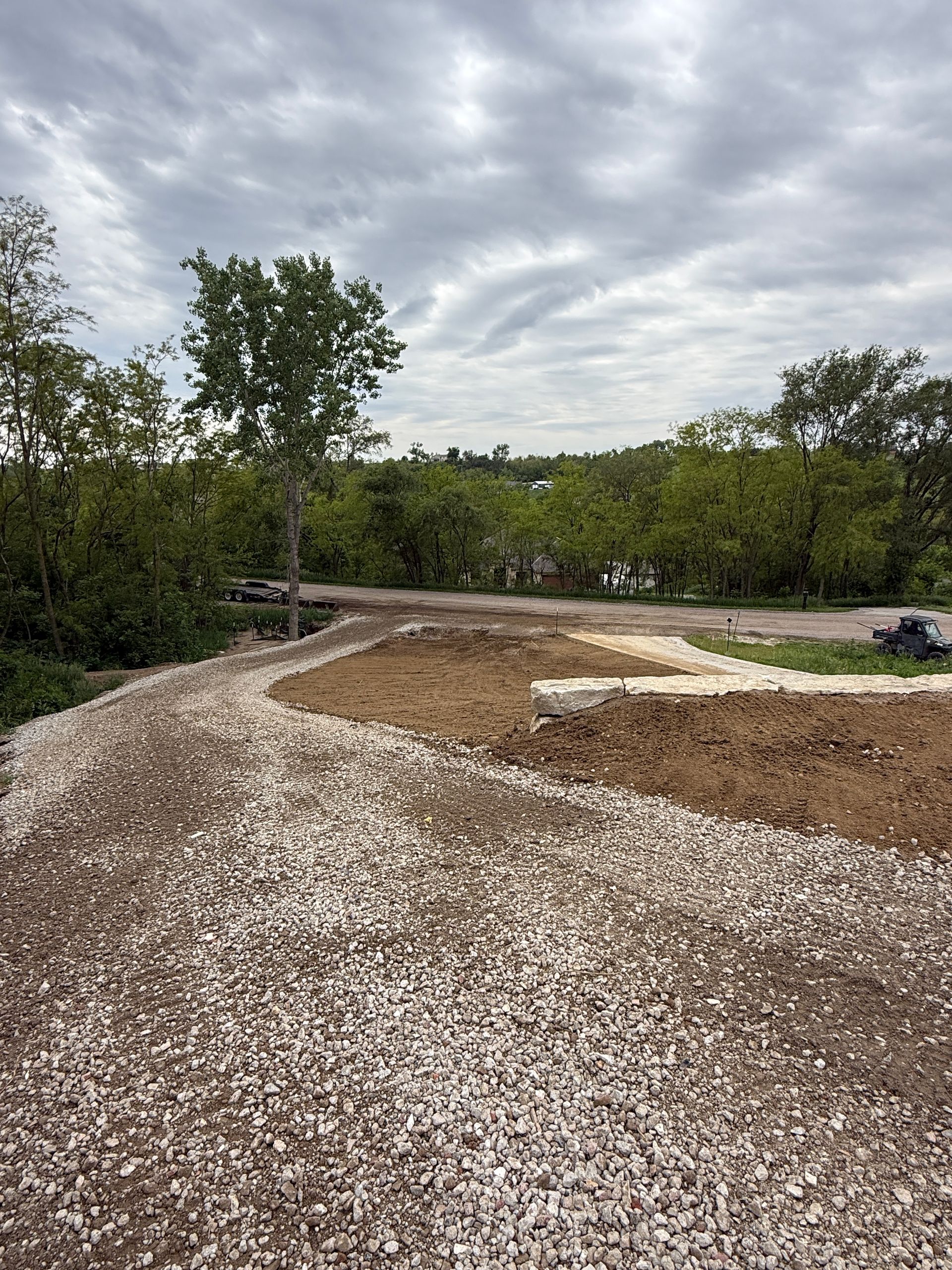 Gravel path curving past a cleared dirt lot with trees under a cloudy sky