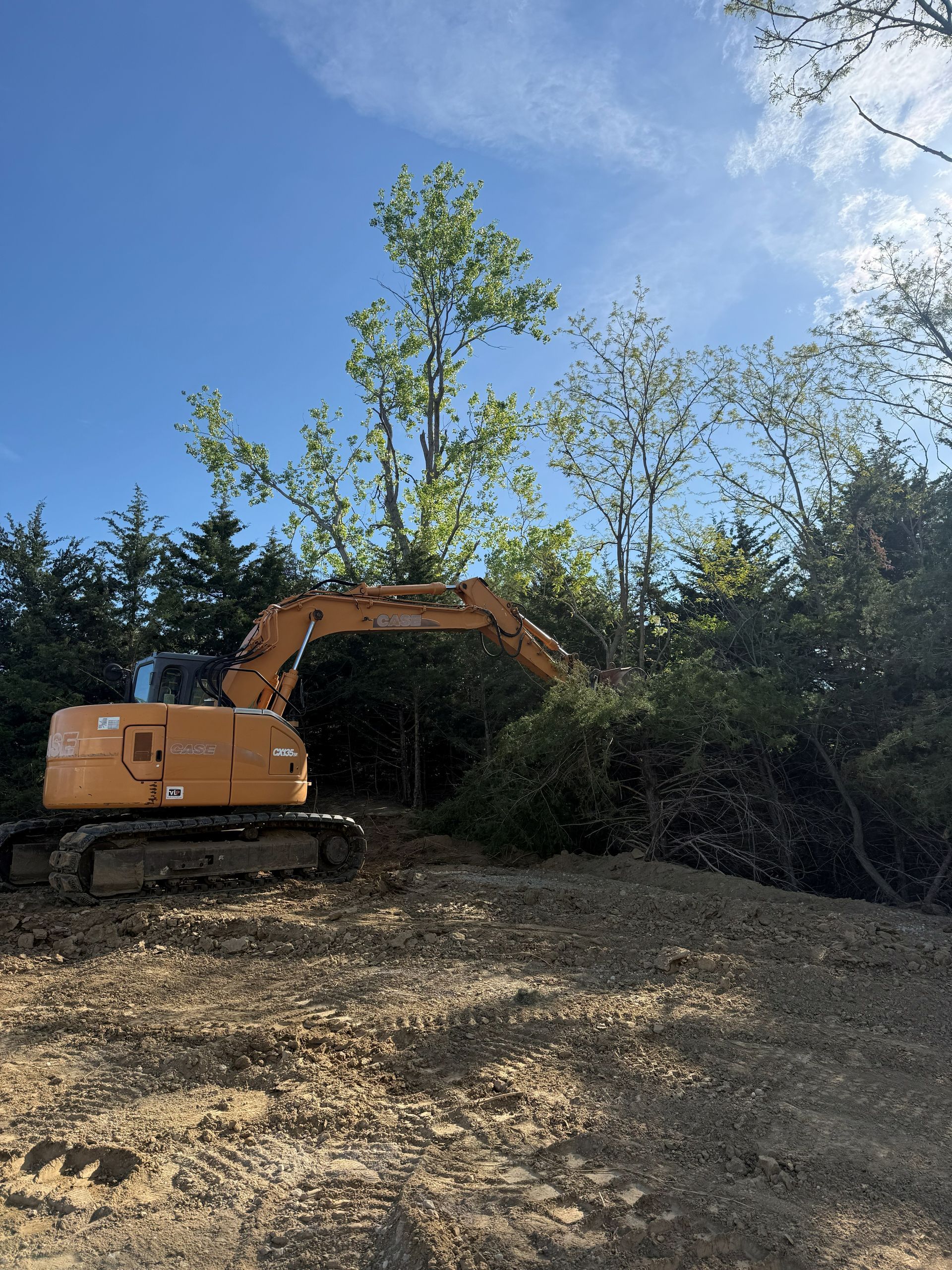 Grassy field with construction materials, equipment, and trees against a bright blue sky.