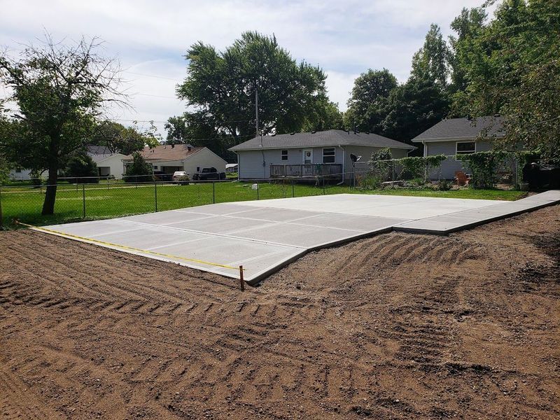 Newly poured concrete pad, gray, in a grassy residential area. Dirt surrounds the edges.