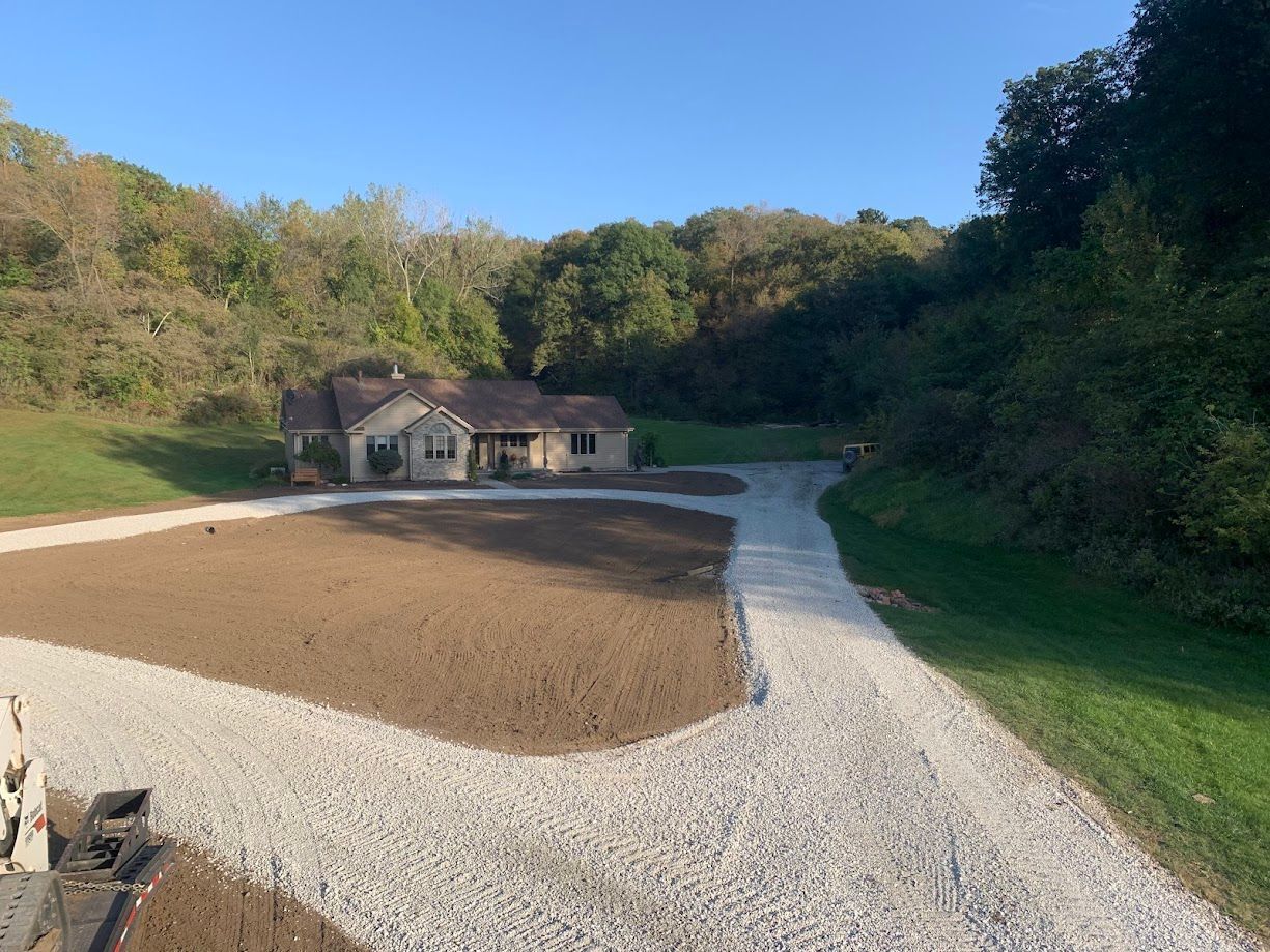 Gravel driveway leads to a small house nestled in a hilly, wooded area, with freshly graded dirt in front.