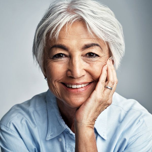 Smiling woman with short white hair resting her face on her hand, wearing a light blue shirt against a gray background