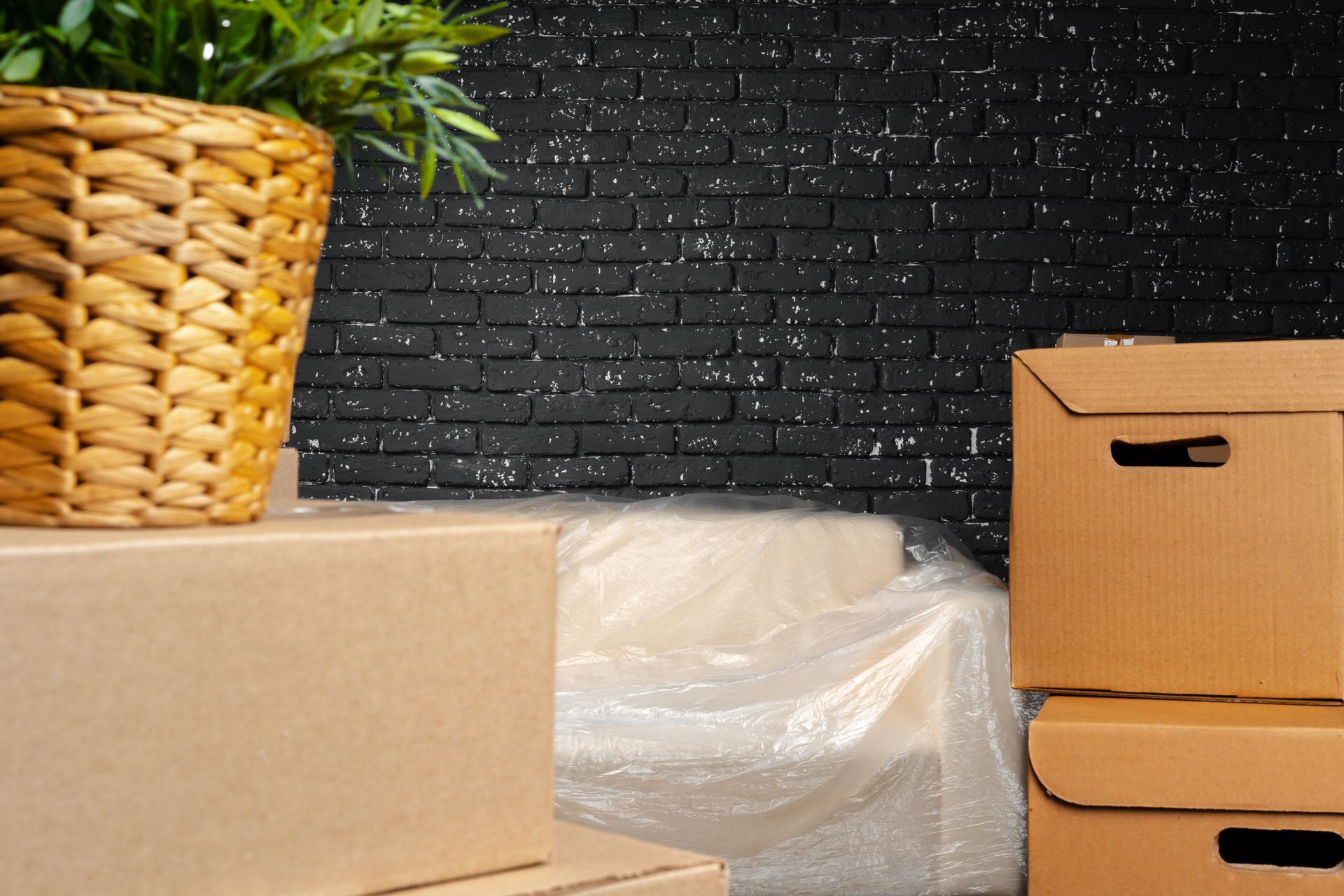 Cardboard boxes, potted plant, and couch with plastic covering against a black brick wall