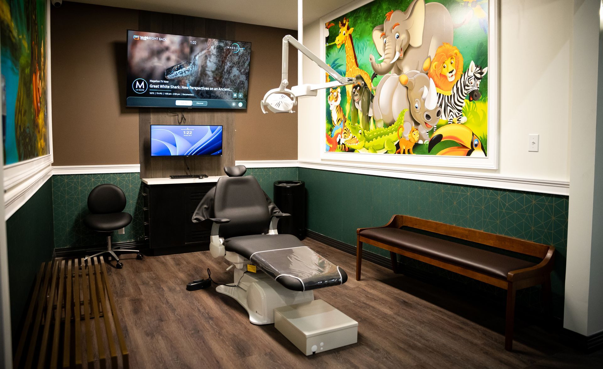Bright, playful dental exam room with a chair, overhead light, TV, and jungle mural on the wall.