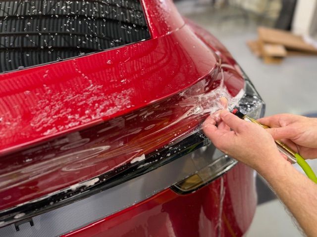 Hands wrapping clear protective film around the red rear bumper of a car