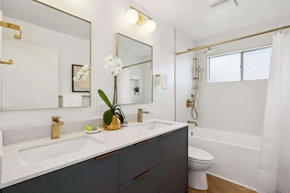 Modern bathroom with gold accents, double vanity, white walls, tub, and window.