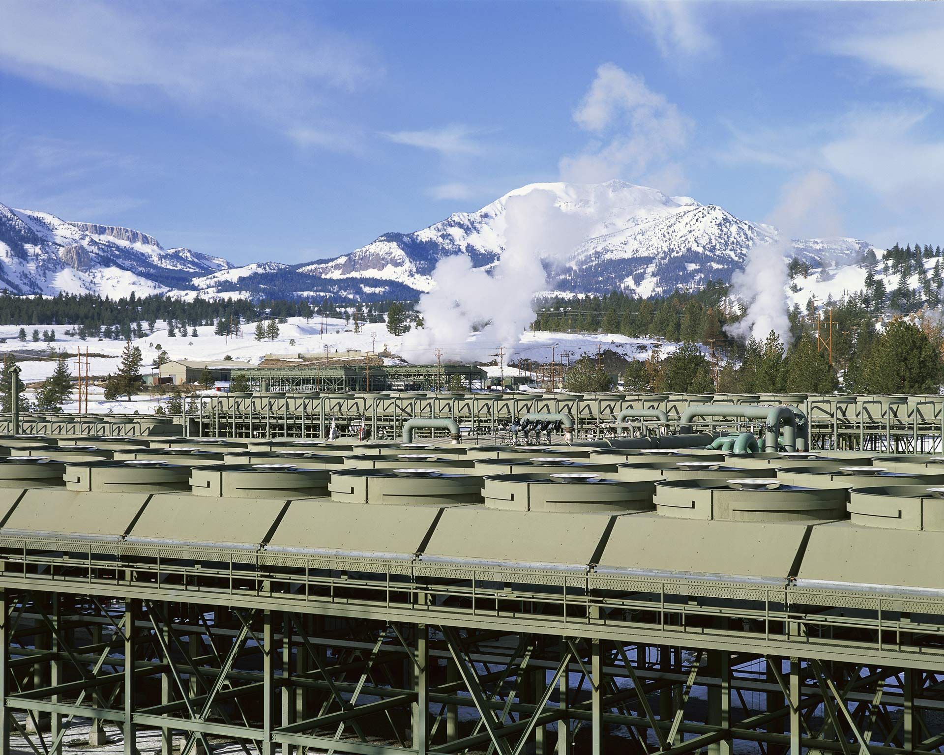 Snowy mountain landscape with steaming industrial structures in the foreground under a blue sky