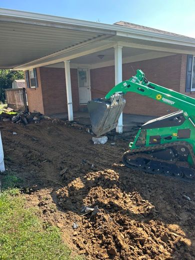 Green mini excavator removing dirt beside a brick house with a covered porch.