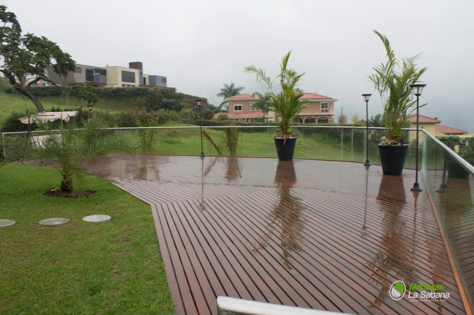 Patio trasero lluvioso con terraza de madera, macetas, césped y piscina más allá de la barandilla de cristal.