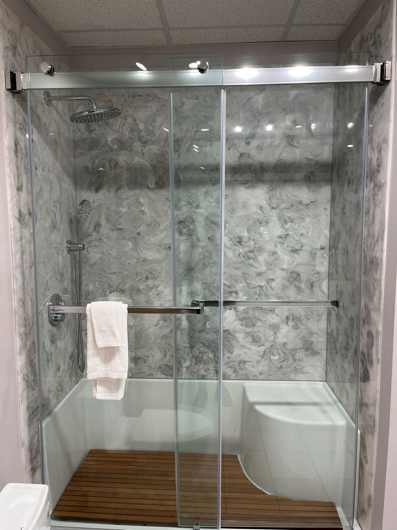 Glass shower stall with gray stone walls, wood floor mat, and a towel on the grab bar.