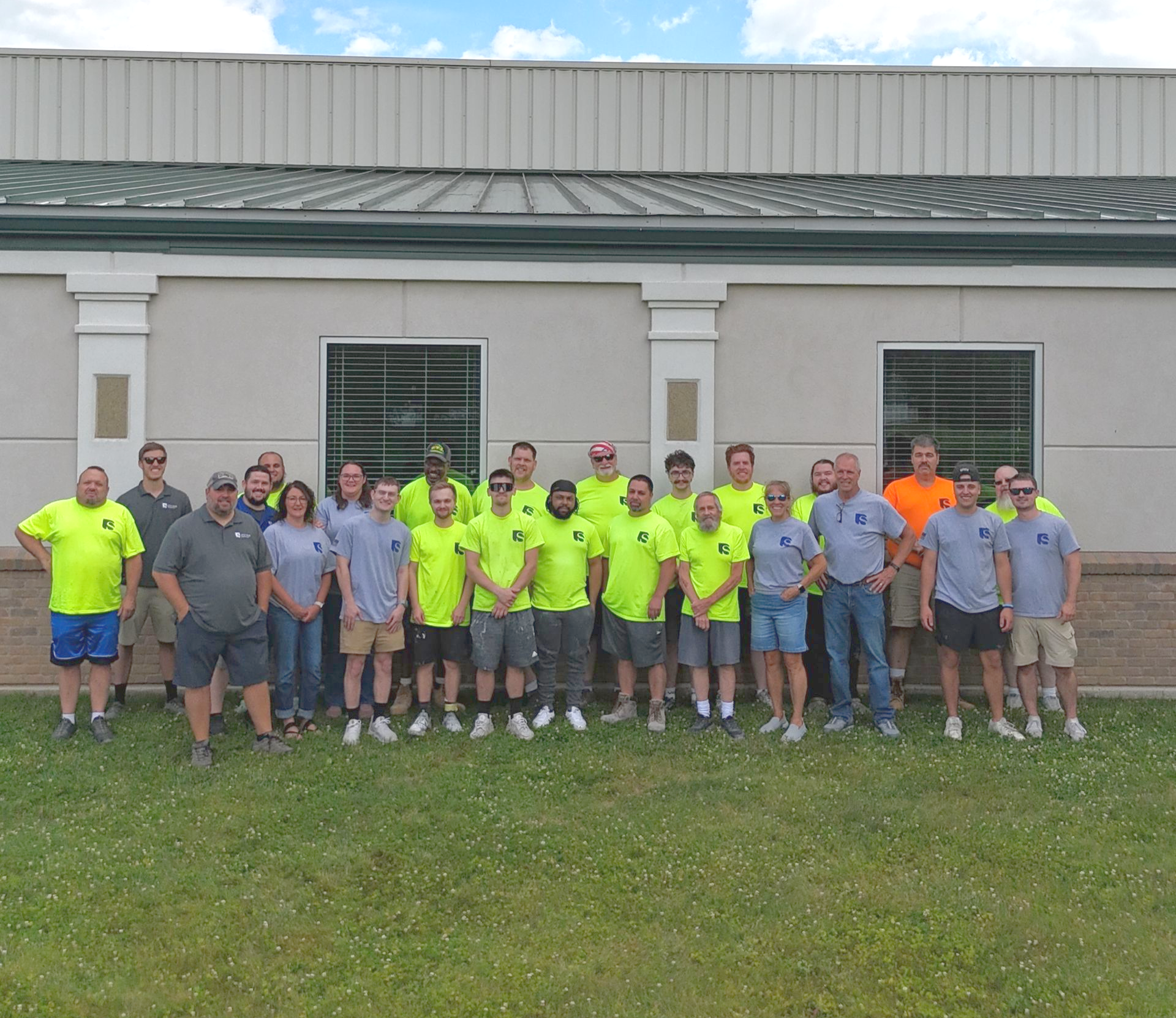 Group of employees in neon shirts posing outside a building with windows and a metal roof.