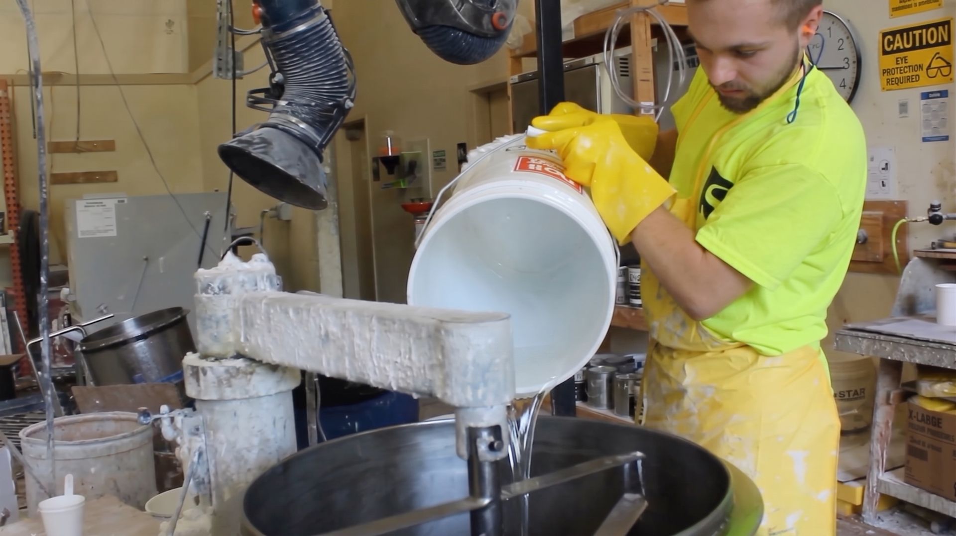 Worker in yellow apron and gloves pouring liquid from a white bucket into a large mixing vat.