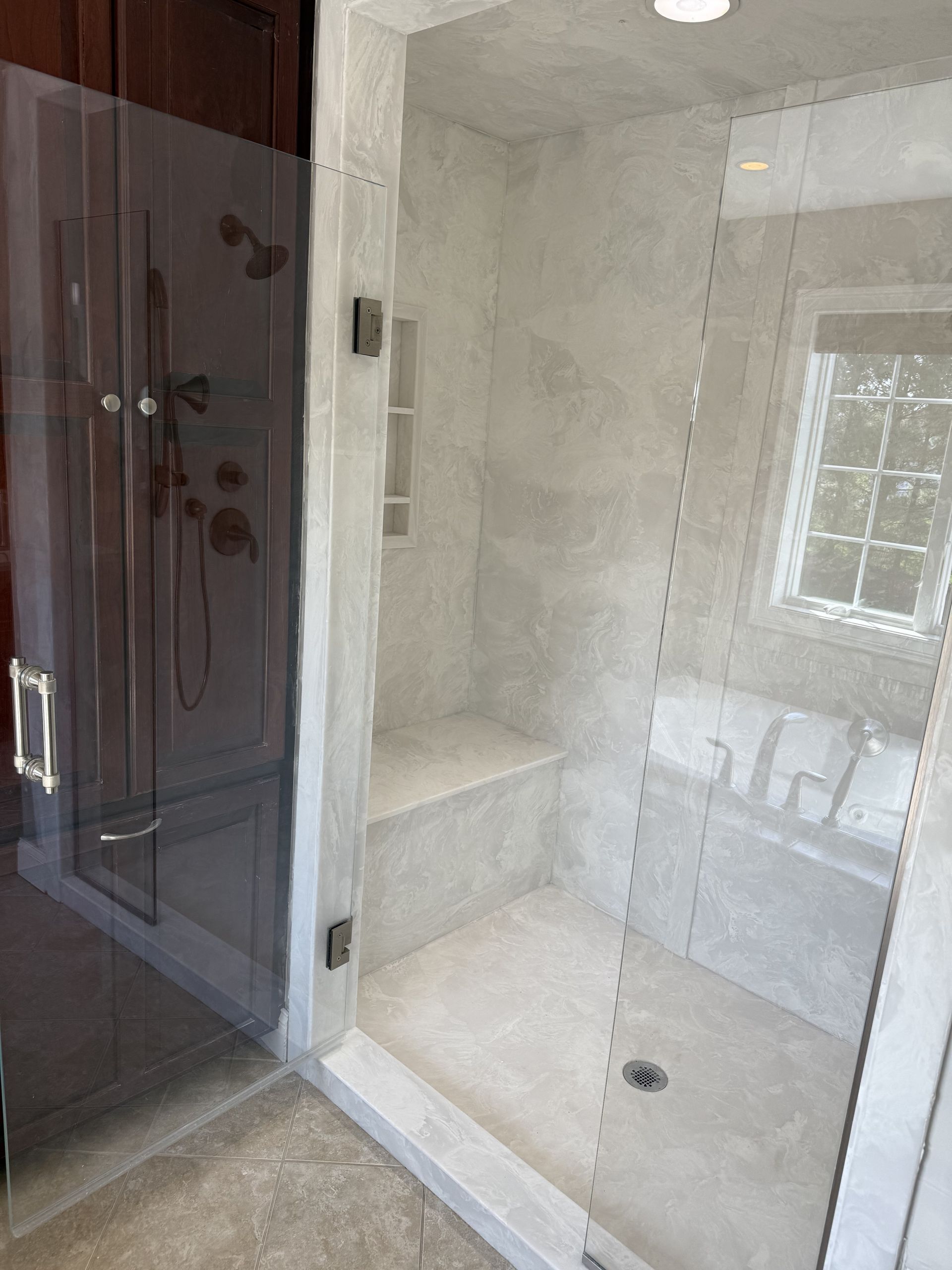 Glass shower stall beside a dark wood cabinet in a tiled bathroom, with a small window inside.