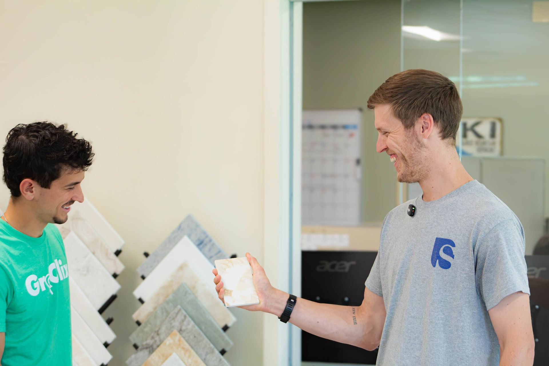 Two men in a showroom smiling and shaking hands by wall displays, one holding a paint sample.