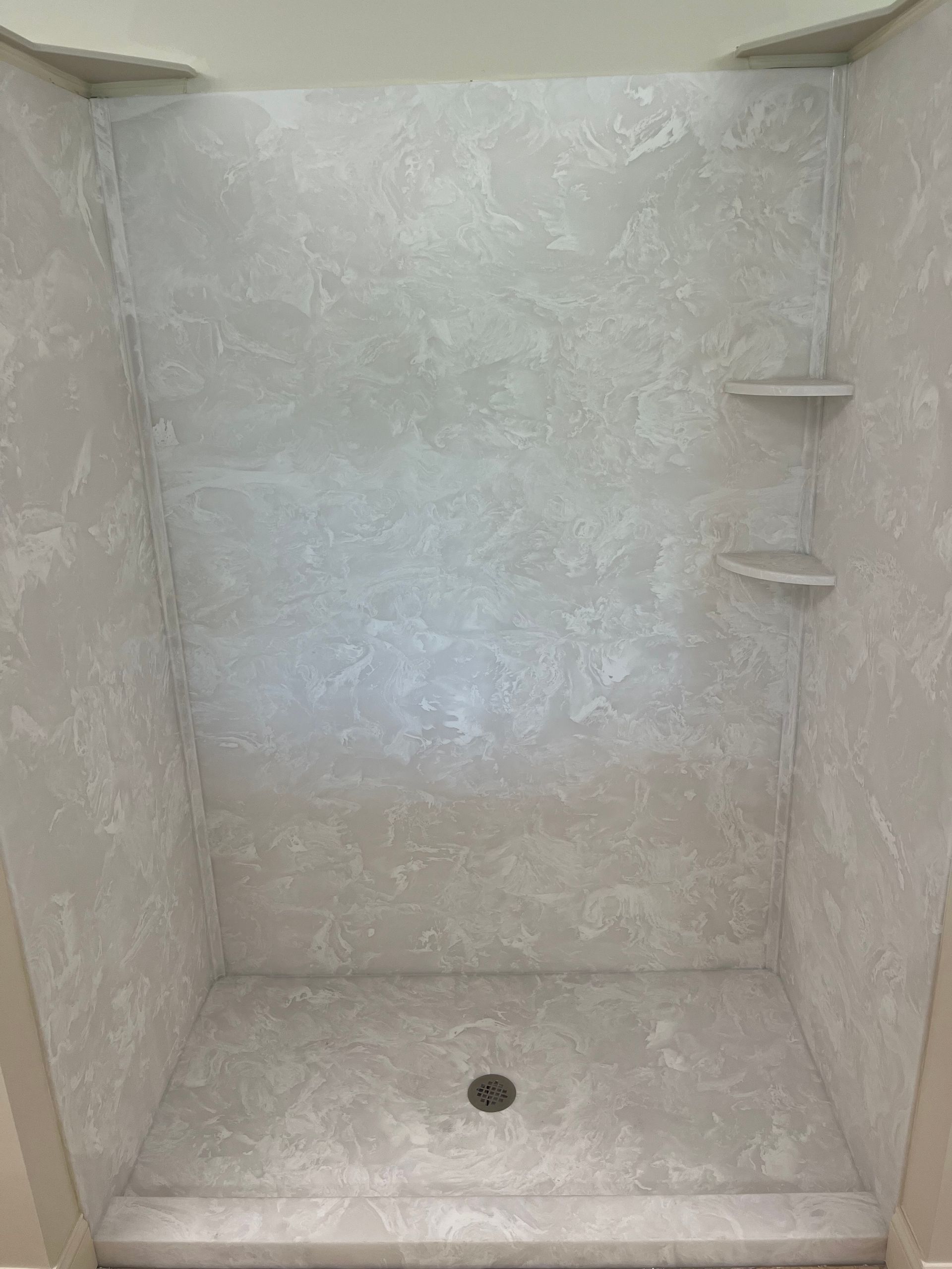 Empty white shower stall with built-in corner shelves and a drain in the floor