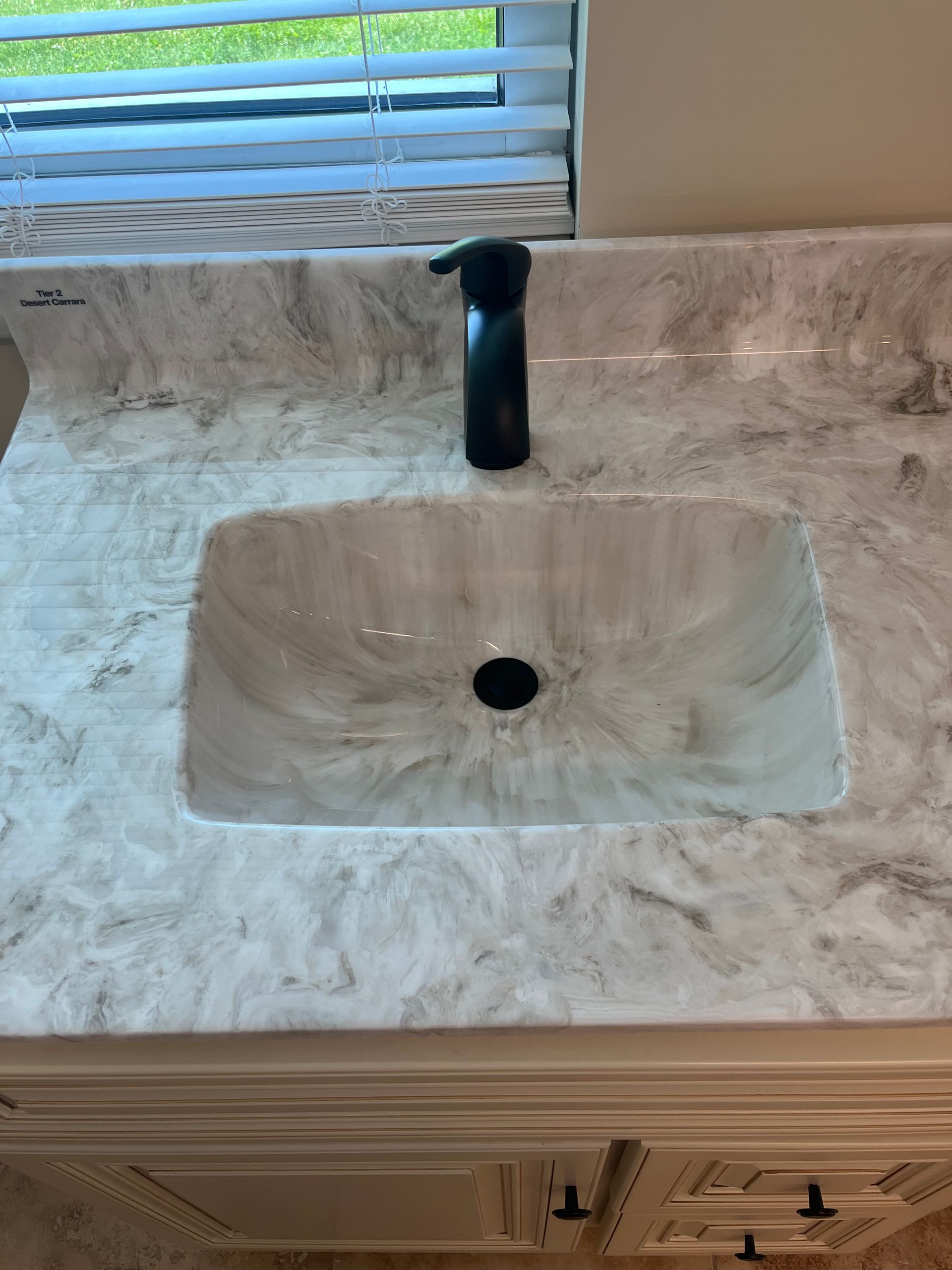 Bathroom sink with black faucet in a marble countertop beneath a window blinds.
