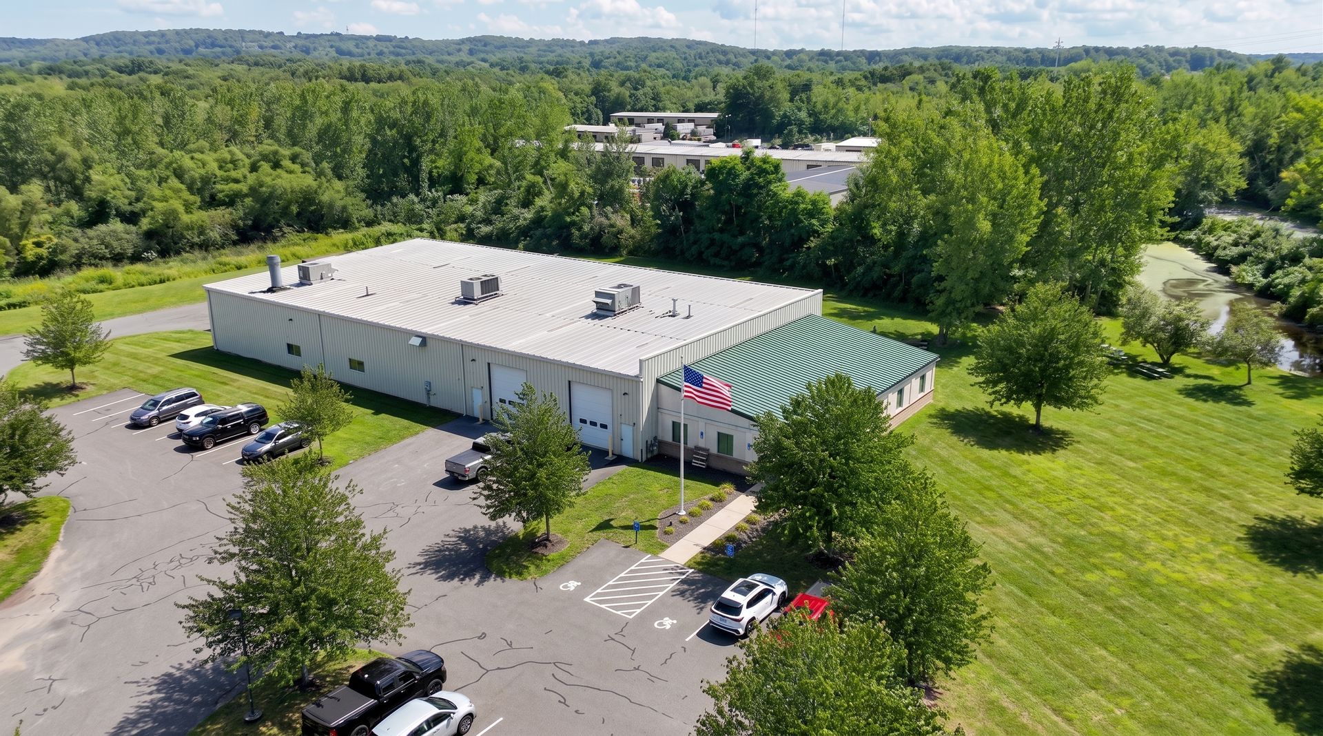 Aerial view of a white industrial building with a green-striped entrance, parking lot, and surrounding trees.
