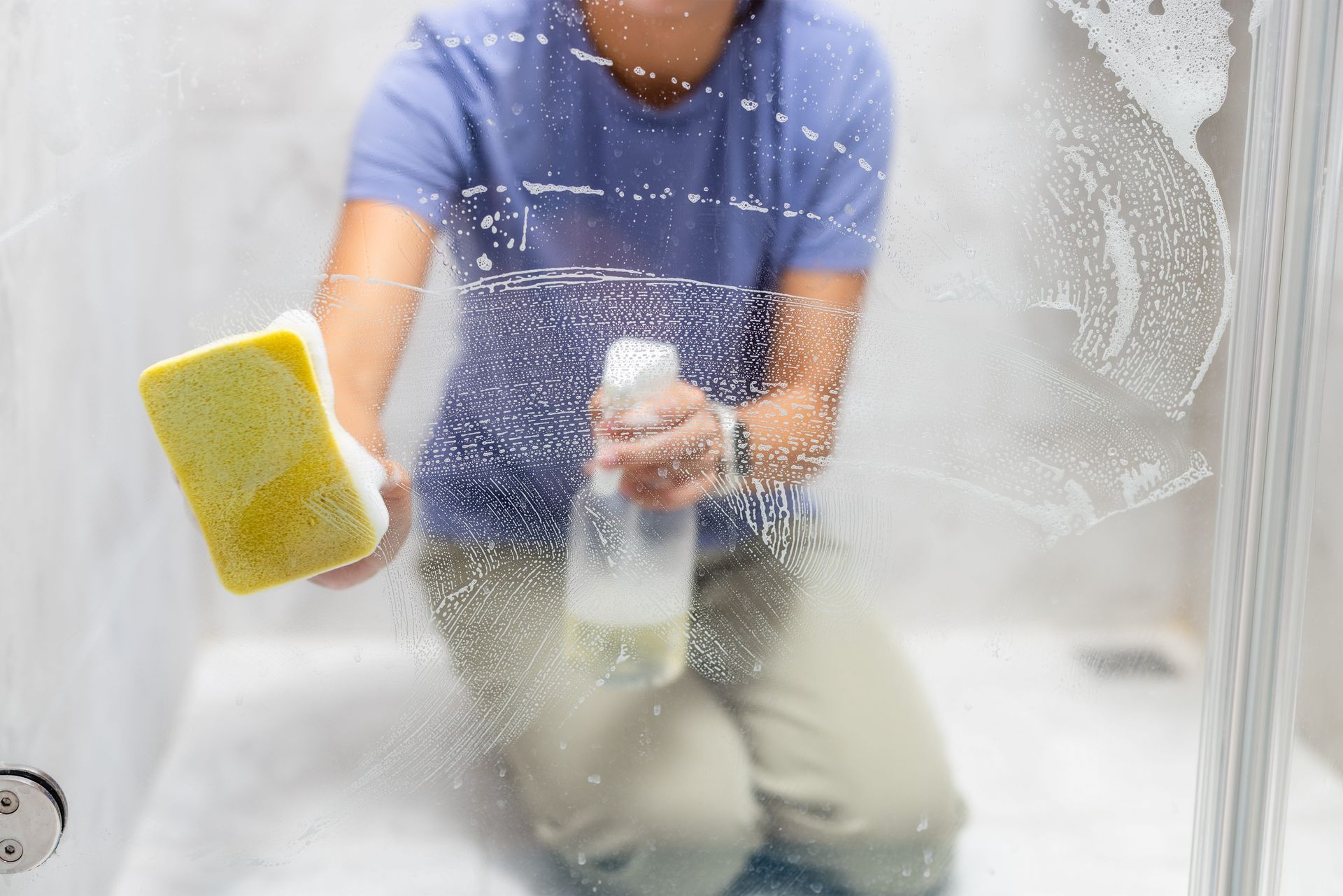 Person cleaning a shower door with a yellow sponge and spray bottle, covered in soap suds