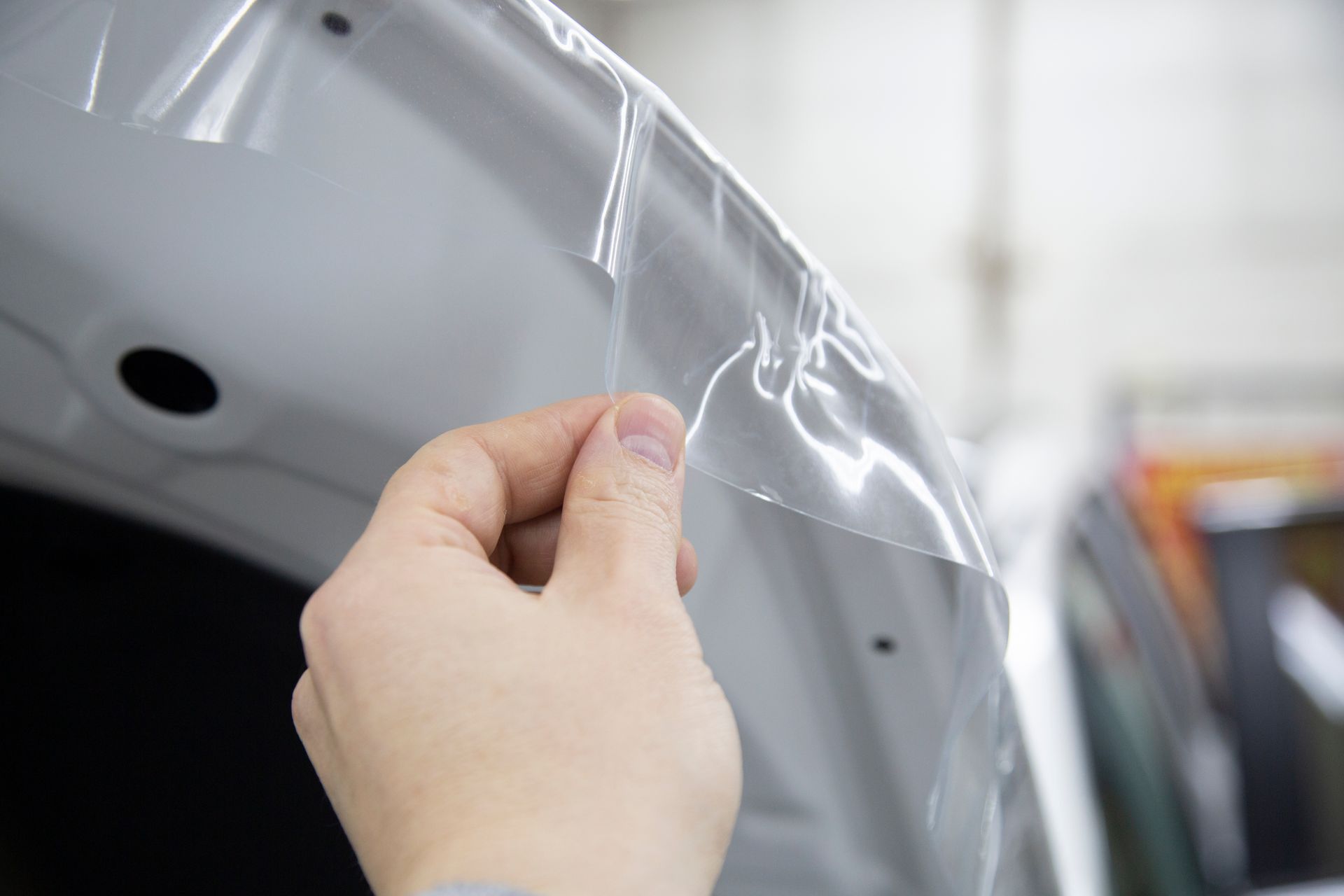 Hand peeling clear protective film from a white car panel in a garage