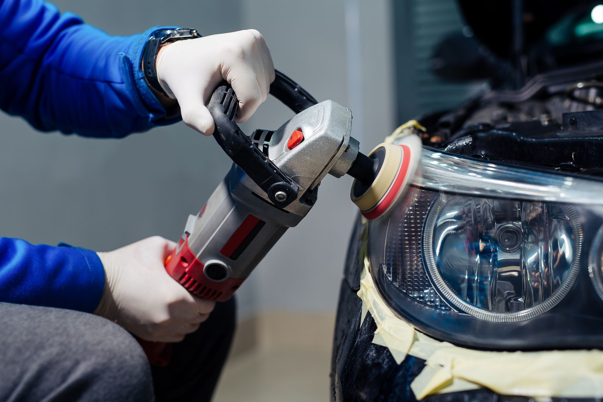 Person polishing a car headlight with a buffer, wearing blue gloves and red sleeves