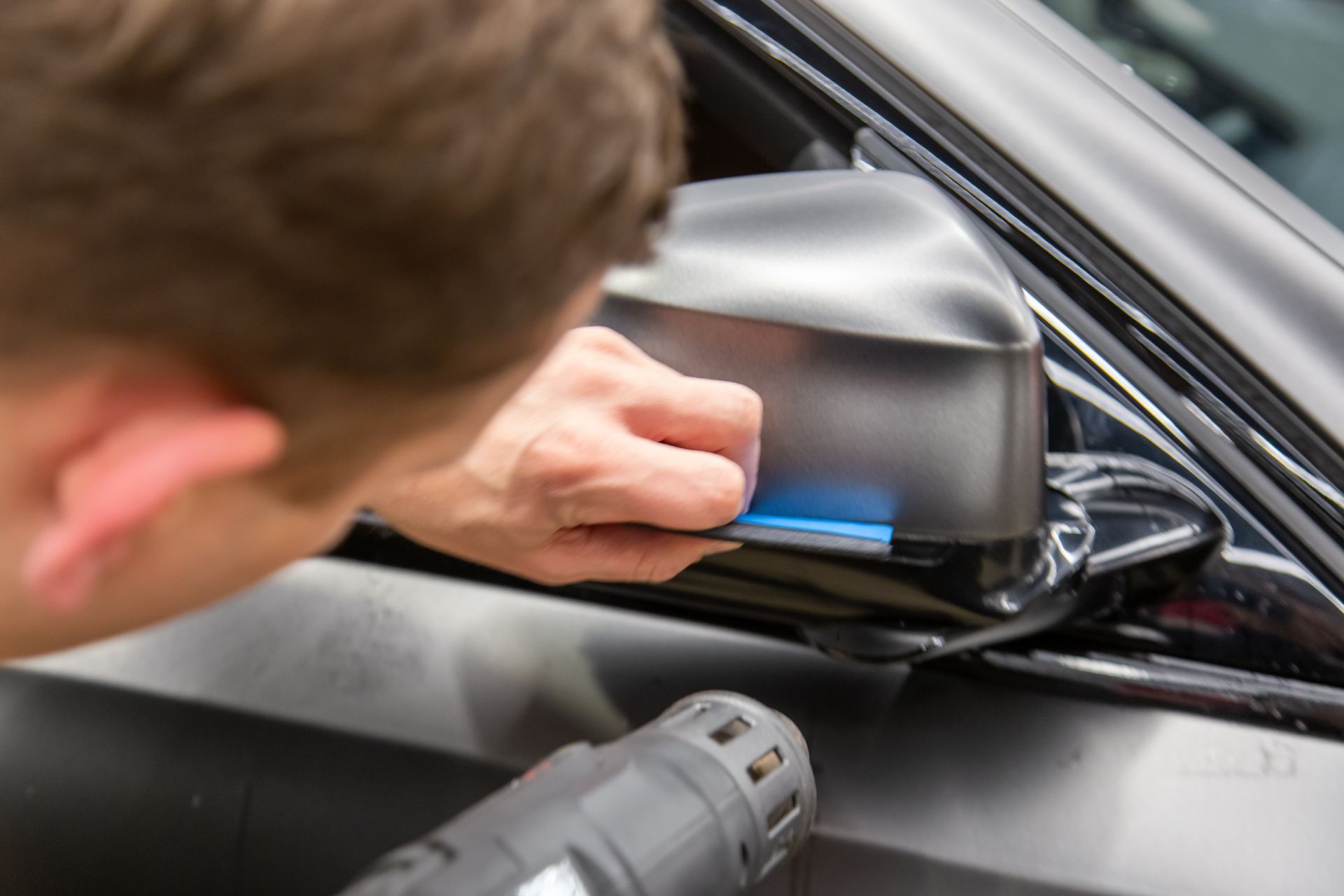 Person polishes a car side mirror with a cloth using a power tool nearby