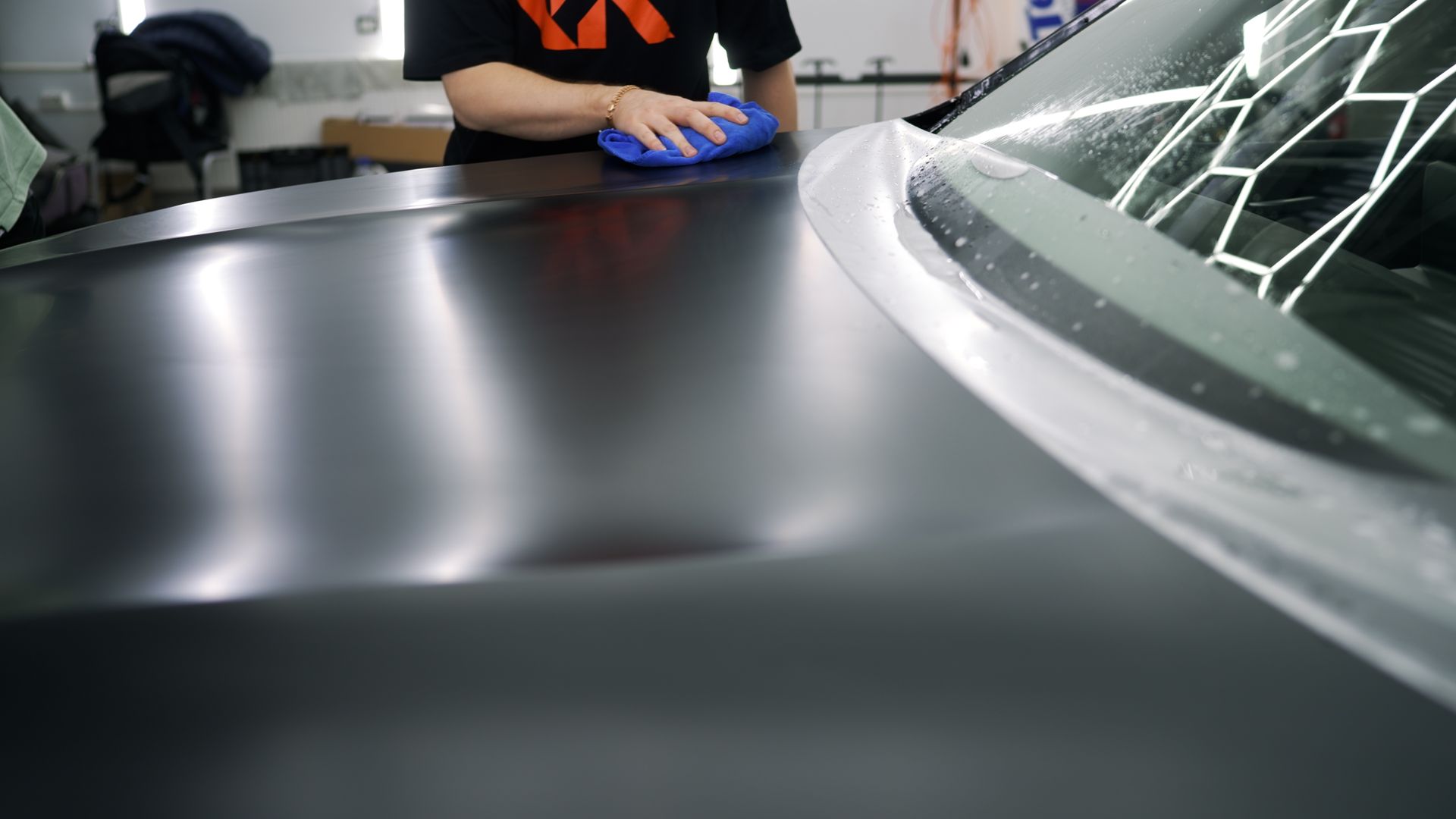 Person in blue gloves smoothing a black car wrap near a windshield in a workshop