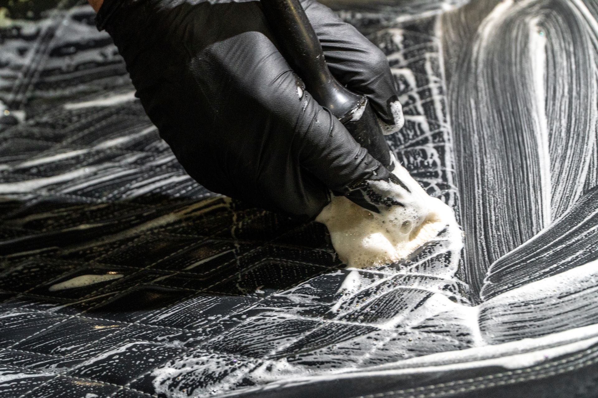 Gloved hand scrubbing a black car hood with soap suds