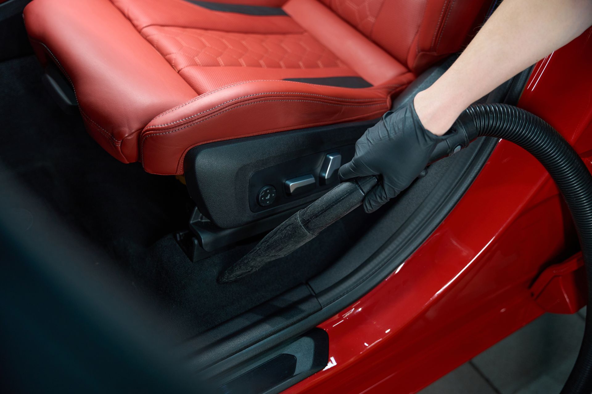 Gloved hand using a vacuum attachment to clean red car seats and interior trim