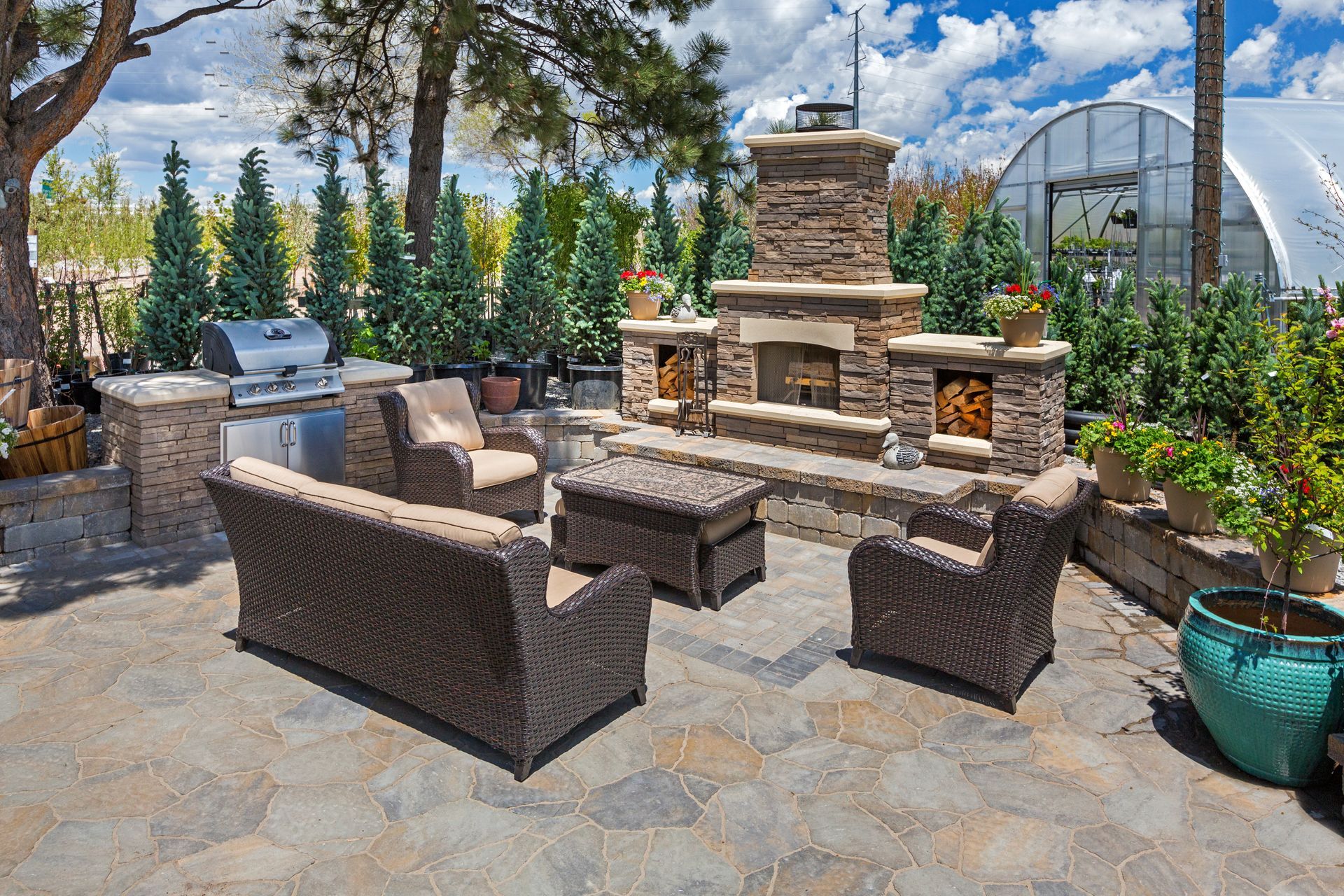 Outdoor stone patio with wicker seating around a brick fireplace, surrounded by trees and potted plants under a blue sky Outdoor stone patio with wicker seating around a brick fireplace, surrounded by trees and potted plants under a blue sky