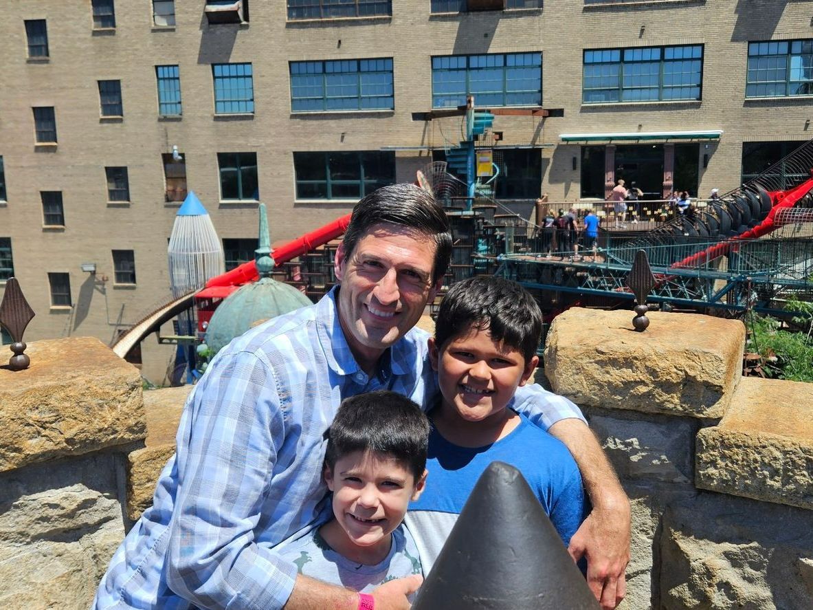 Man and two boys smile, posing together. Outdoor setting with buildings and play equipment visible.