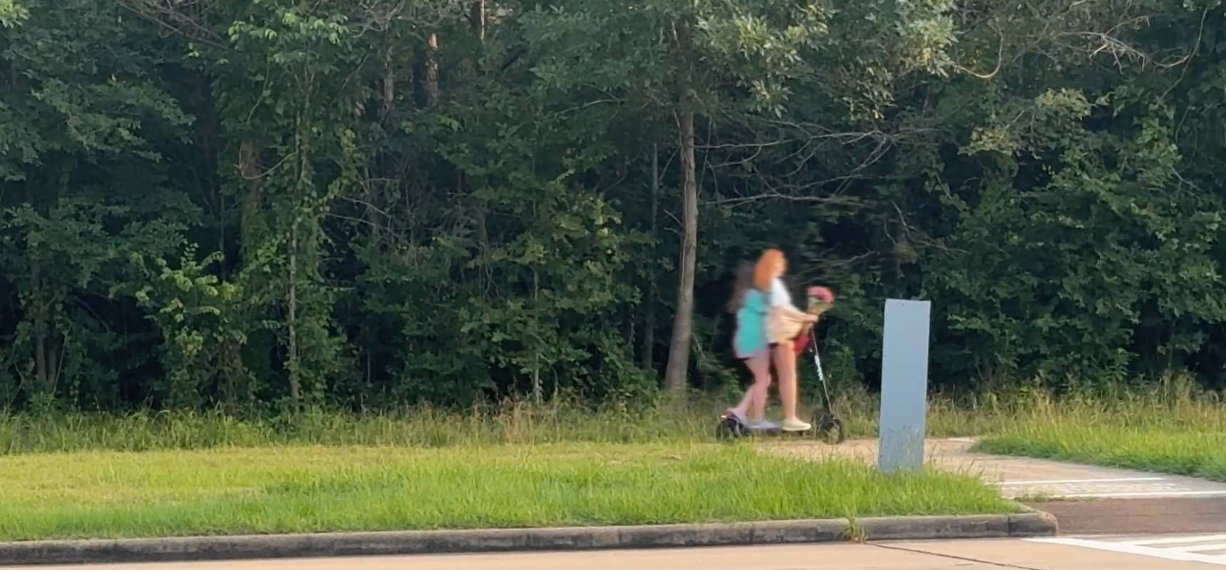 Two children ride an electric scooter on a sidewalk in The Woodlands, Texas.