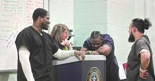 Three people stand at a podium while one person rests their head on it in a classroom setting.