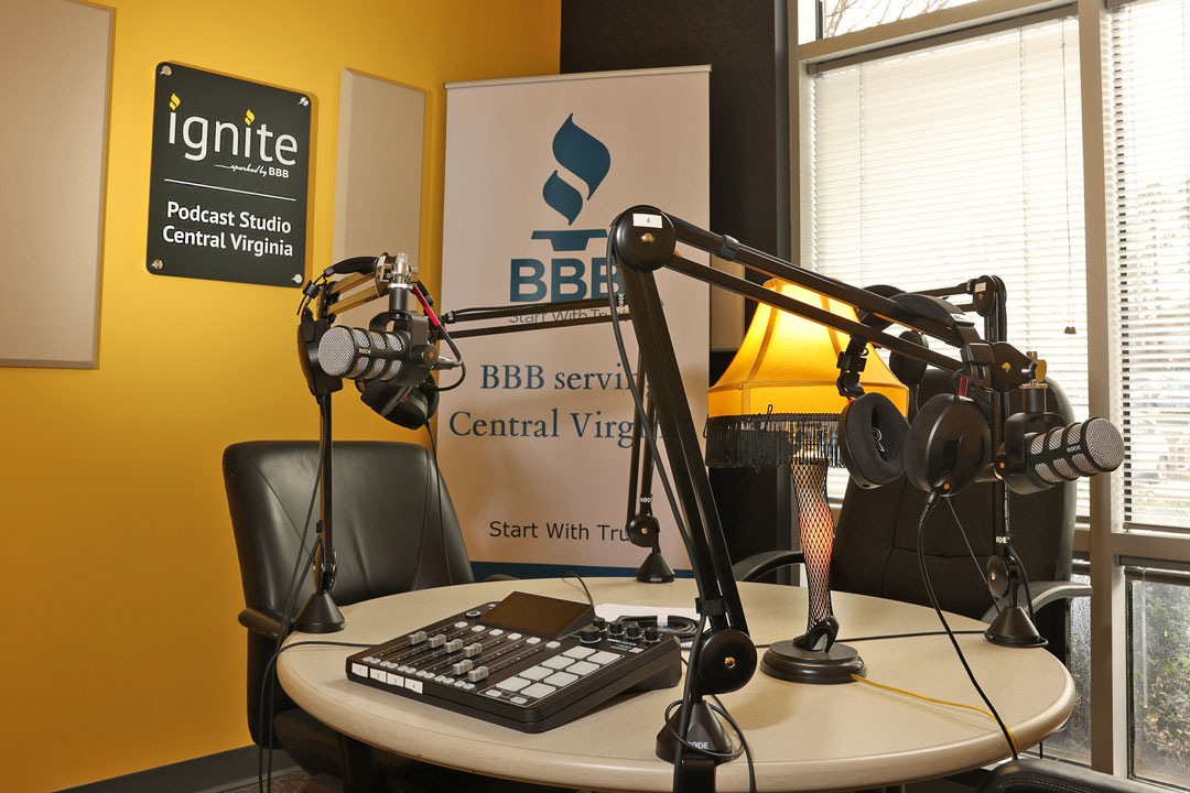 Radio studio with microphones on boom arms, a control board, and a banner.