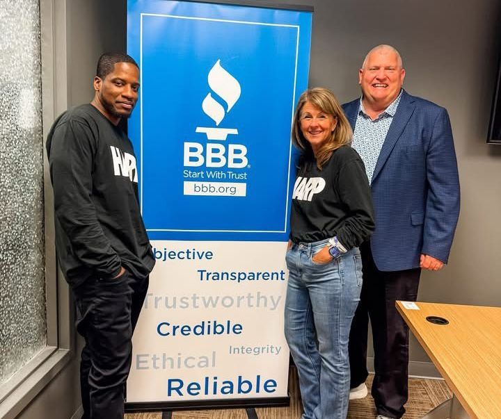 Three people posing beside a BBB sign in an office, with a blue banner and values text visible.
