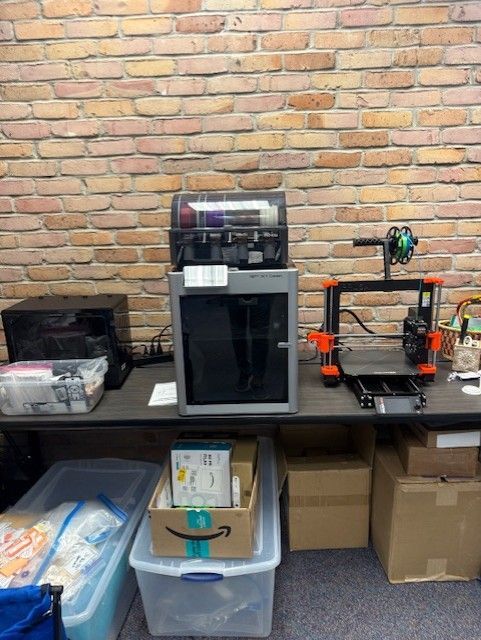 Counter with espresso machine, plastic bins, cardboard boxes, and a small cart against a brick wall.