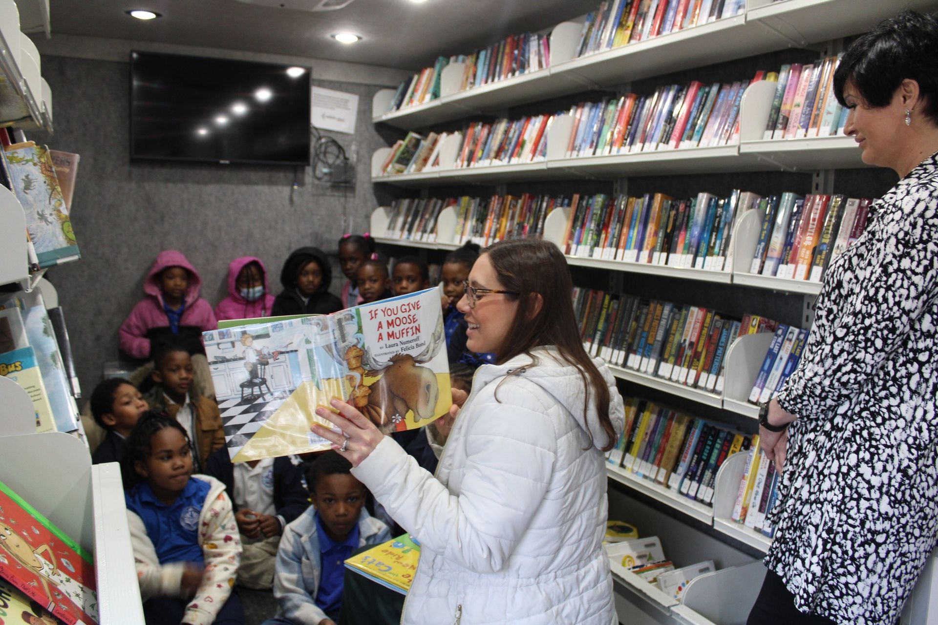Woman reads a book to children in a library, with shelves of books in the background.