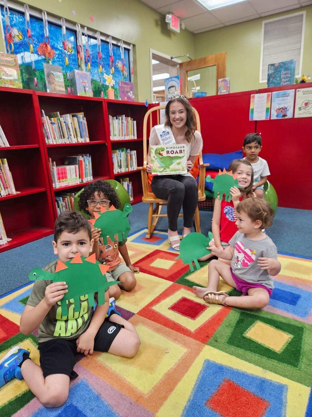 Teacher and children sitting on a colorful classroom rug, holding green paper leaf crafts.
