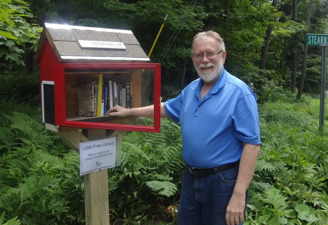 Man in blue shirt pointing to a red little free library in a wooded area